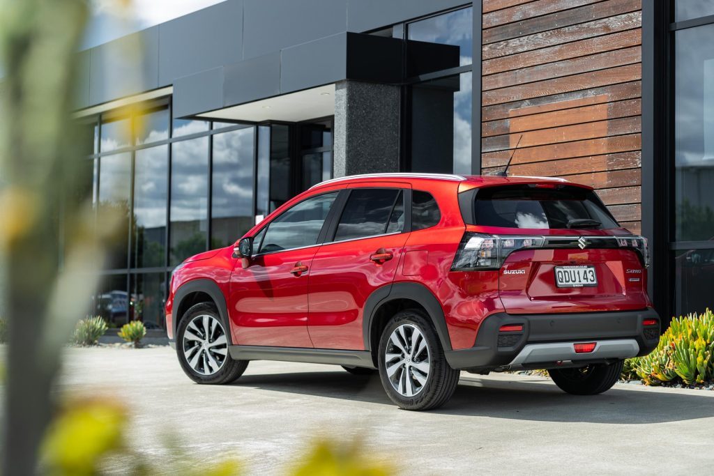 Rear quarter profile of the Suzuki S-Cross Hybrid in red, parked next to office
