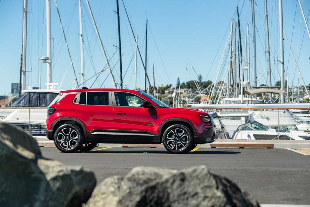 Side profile of the Jeep Avenger, pitcured in red, parked behind rocks at an Auckland marina