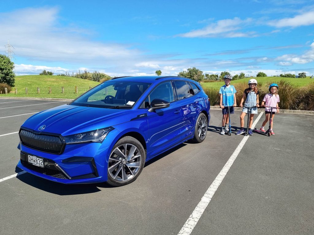Blue Skoda Enyaq, with kids lined up on scooters